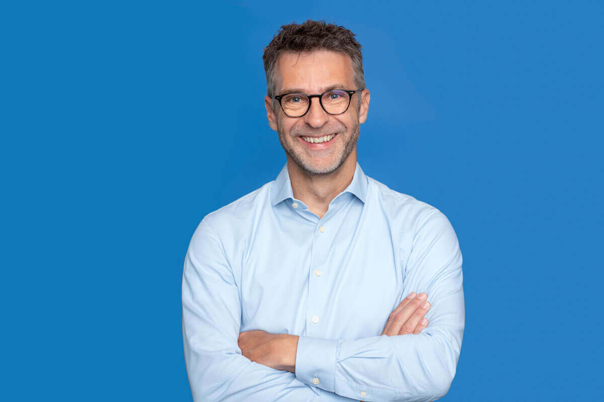 Mann vor blauem Hintergrund, schaut lächelnd in die Kamera