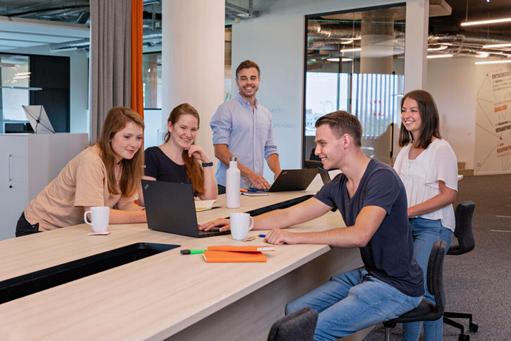 5 Werkstudenten sitzen an einem großen Tisch. Um sie herum stehen Kaffeetassen und Notziblöcke. Die Blicke sind auf den vordersten Laptop gerichtet.